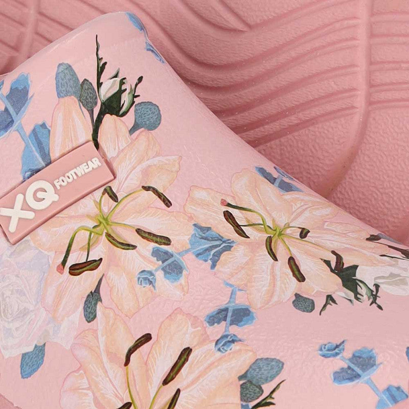 Close-up van de XQ Tuinklompen Dames met Bloemenprint- Instapmodel & Lichtgewicht in roze met een lelies en blauwe bloemenprint, waterafstotend design en XQ Footwear label op een gestructureerde roze achtergrond.