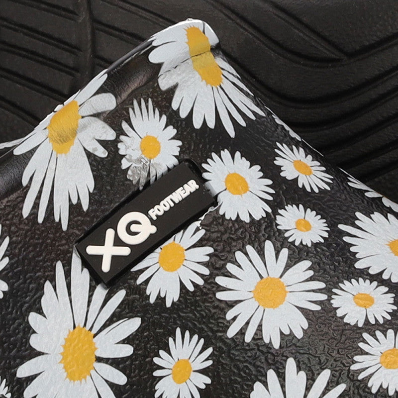 Een close-up van de XQ Tuinklompen Dames met Margrietjesprint, een lichtgewicht EVA slip-on met een zwart oppervlak met witte madeliefjes, het "XQ FOOTWEAR" label in wit en een zwarte rubberen zool zichtbaar op de achtergrond.