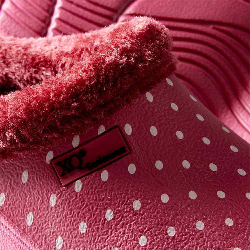 Close-up van de XQ Tuinklompen Dames Met Bontje en Stippenprint - Instapmodel & Lichtgewicht: een roze slipper met witte stippen, pluizige roze voering en label aan de zijkant. De achtergrond laat meer zien van het getextureerde, warme gevoerde klompenmateriaal.