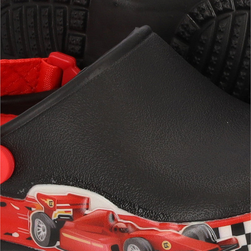 Een close-up van de XQ Tuinklompen Jongens, een zwarte klompschoen voor kinderen met LED-lampje, met een Formule 1-print raceauto op de zijkant, rode band en zwarte zolen met textuur op de achtergrond.