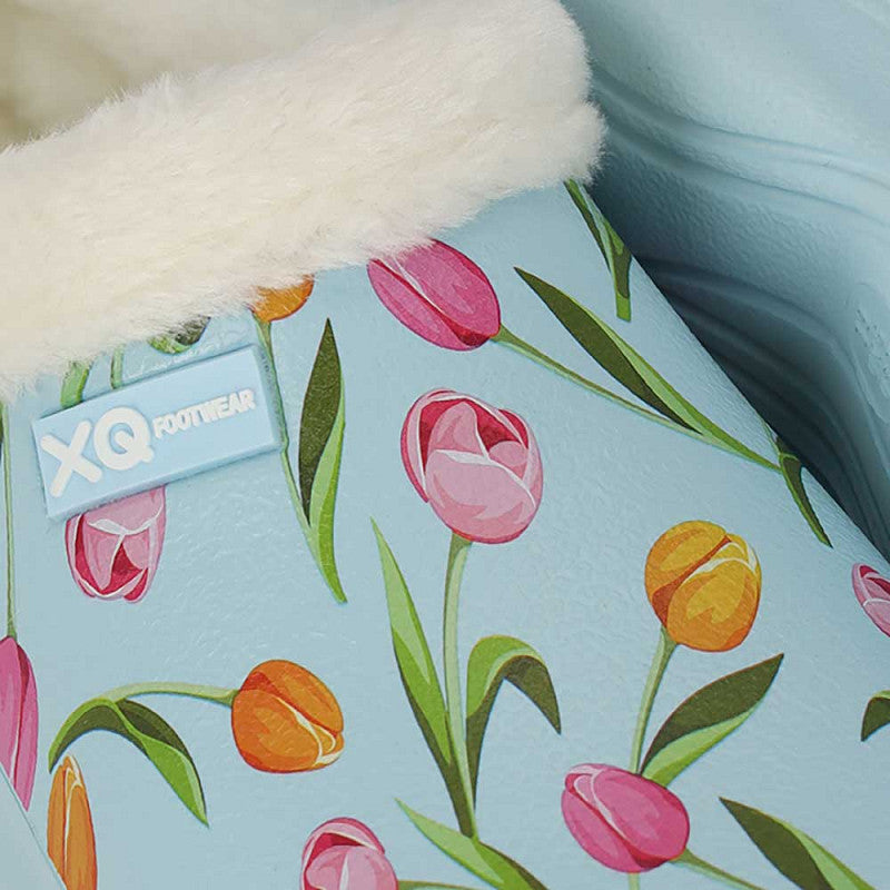 Een close-up van de XQ Tuinklompen Dames - Met Bontje & Tulpenprint: lichtblauwe damesklompen met een pluizige witte voering, een levendige tulpenprint en een XQ Footwear-label.