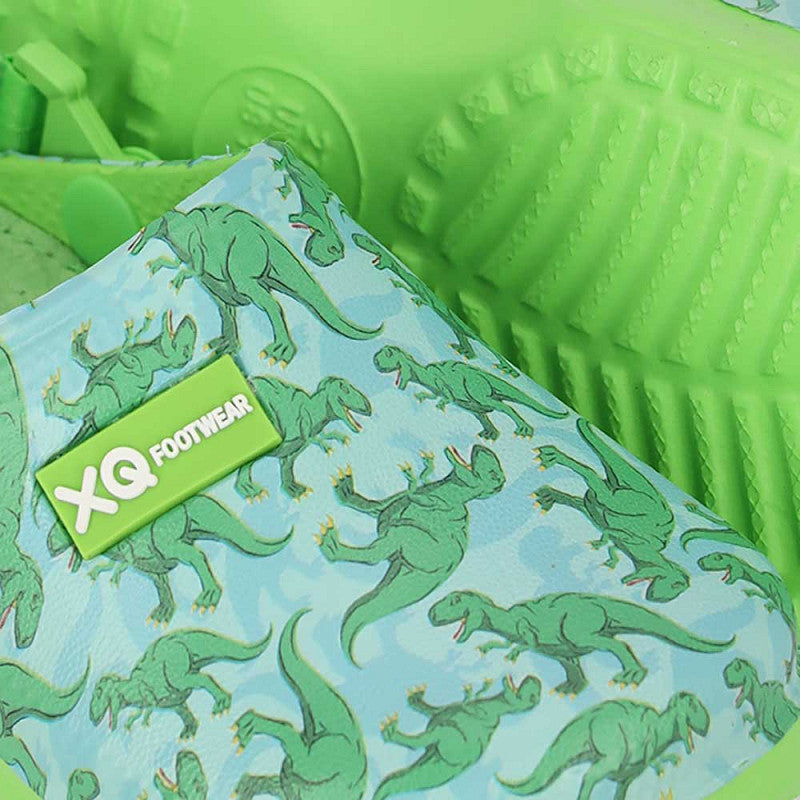 Een close-up van XQ Tuinklompen jongens - Klittenband & Dinoprint, met lichtblauwe stof met een groen dinosauruspatroon, felgroene antislip zool en een groen label met witte XQ FOOTWEAR tekst.