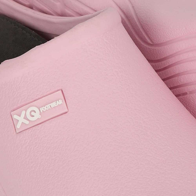 Close-up van XQ Tuinklompen Dames Licht Roze - Instapmodel & Lichtgewicht met een gestructureerd lichtroze oppervlak en wit XQ FOOTWEAR-logo op de zijkant. Op de achtergrond is nog een bijpassende tuinklomp te zien.