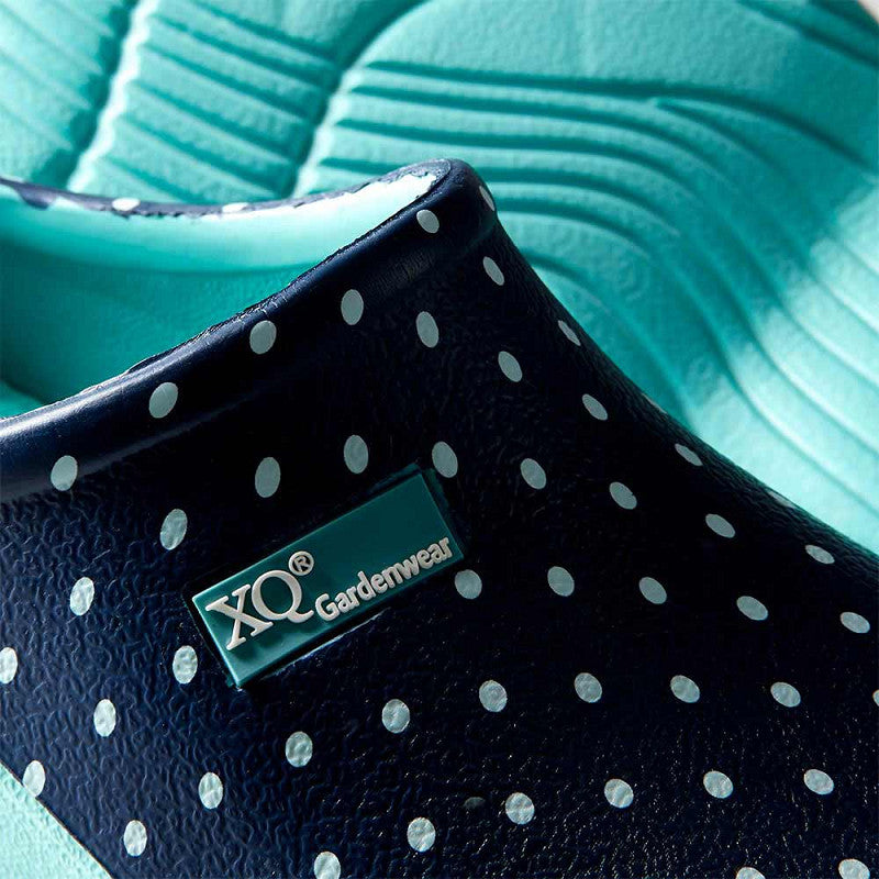 Close-up van XQ Tuinklompen Dames - Lichtgewicht & Stippenprint in marineblauw met witte stippen, met een XQ Gardenwear-label, gemaakt van lichtgewicht EVA tegen een turquoise getextureerde achtergrond.