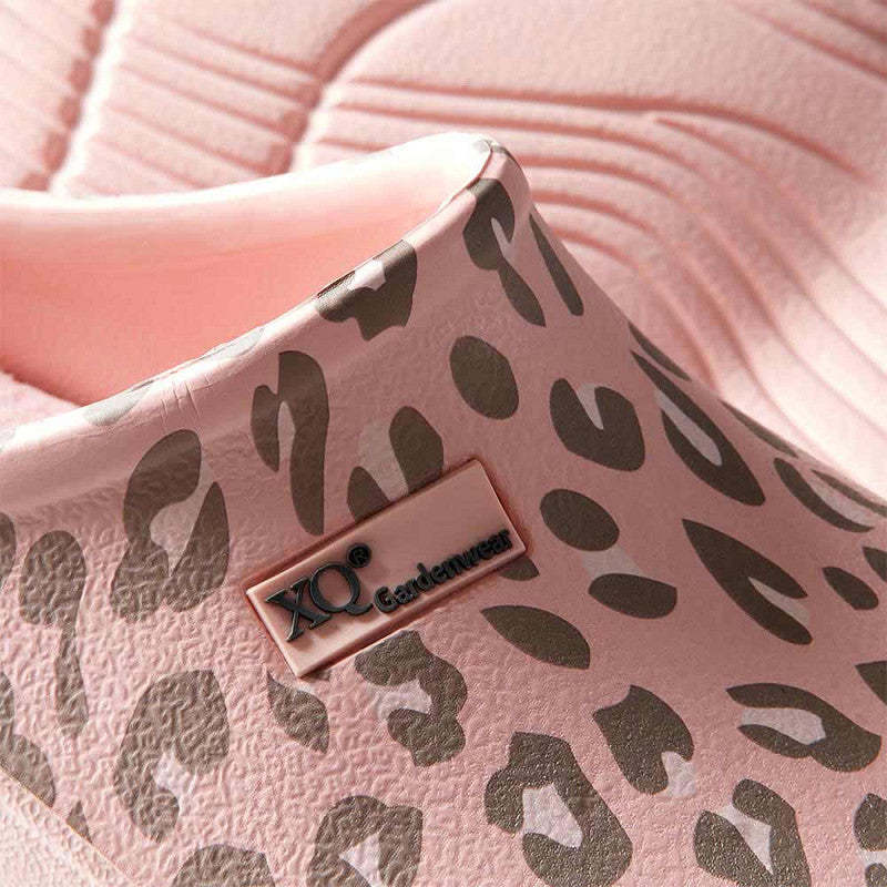 Close-up van de XQ Tuinklompen Dames met Luipaardprint roze-instapmodel, lichtgewicht, waterafstotend en met grijze luipaardprint. Op de zijkant zit een XQ Gardenwear-label; op de achtergrond een getextureerd roze oppervlak.