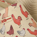 Een close-up van beige XQ Tuinklompen Dames met Dierenprint - Kippen toont rode en witte kippenmotieven en een label met 'XQ FOOTWEAR' in rood, tegen een subtiel golvende achtergrond die de leuke tuinklompenstijl complementeert.