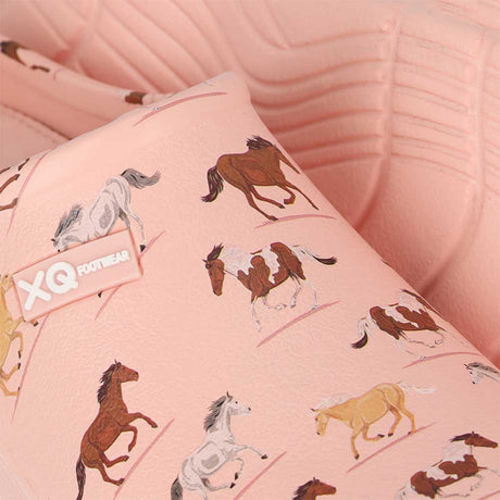 Close-up van de met Dierenprint - Paarden XQ Footwear sandaal in roze, met rennende paarden in bruin, wit en bruin. De waterafstotende tuinklompen liggen op een getextureerde roze achtergrond.